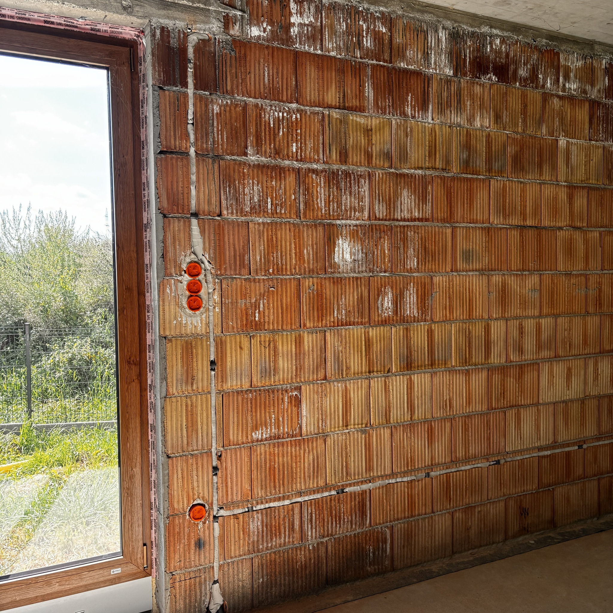 Ściana z pustaków z instalacją elektryczną w trakcie montażu, widoczne puszki i przewody. Okno z widokiem na zieleń w tle. Surowy stan wykończenia.