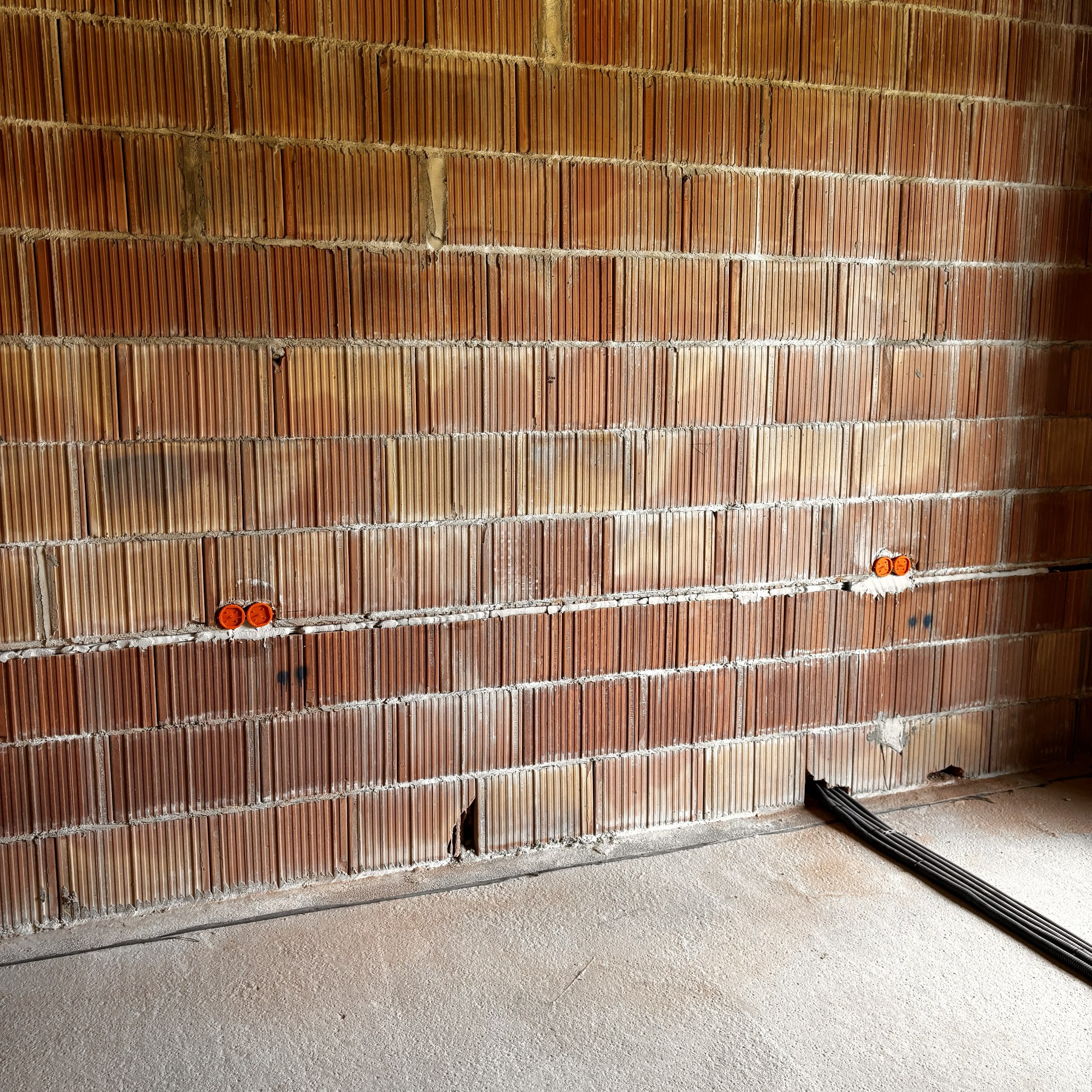 Czerwona ściana z pustaków z widocznymi puszkami elektrycznymi i kablami na podłodze. Etap prac instalacyjnych w surowym wnętrzu.