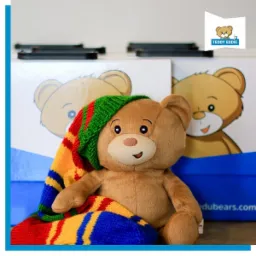 Pluszowy miś z uśmiechem, ubrany w kolorową, dzierganą czapkę w paski, siedzi na tle pudełek z rysunkami misia i logo 'Teddy Eddie'.