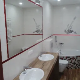 Nowoczesna łazienka z dwoma umywalkami na drewnianym blacie, duże lustro z czerwoną ramą, białe płytki z motywem gitary i skrzypiec, mozaika na obudowie wanny.