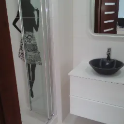 Nowoczesna łazienka z kabiną prysznicową z grafiką kobiety, czarną umywalką na białej szafce i lustrem z widocznym fragmentem ciemnych drzwi.