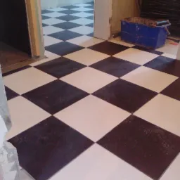 Ułożona podłoga z płytek w czarno-białą szachownicę, widok z góry na świeżo położone płytki, w tle wiadro z narzędziami i otwarte przejście do kolejnego pomieszczenia.