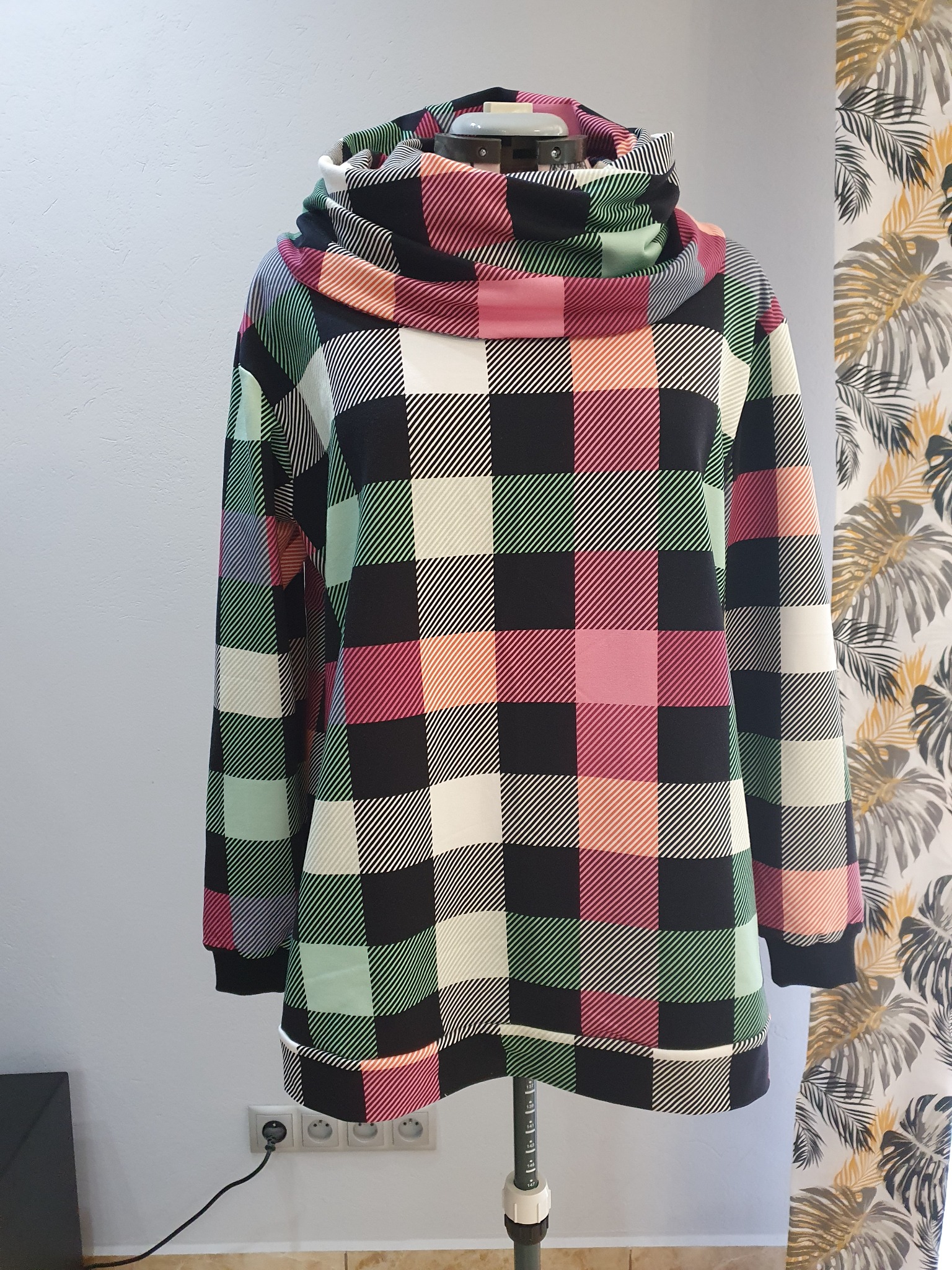 Bluza z obszernym golfem w dużą kratę, uszyta z materiału w odcieniach czerni, bieli, różu, zieleni i pomarańczy, prezentowana na manekinie krawieckim na tle ściany i zasłony w liście.