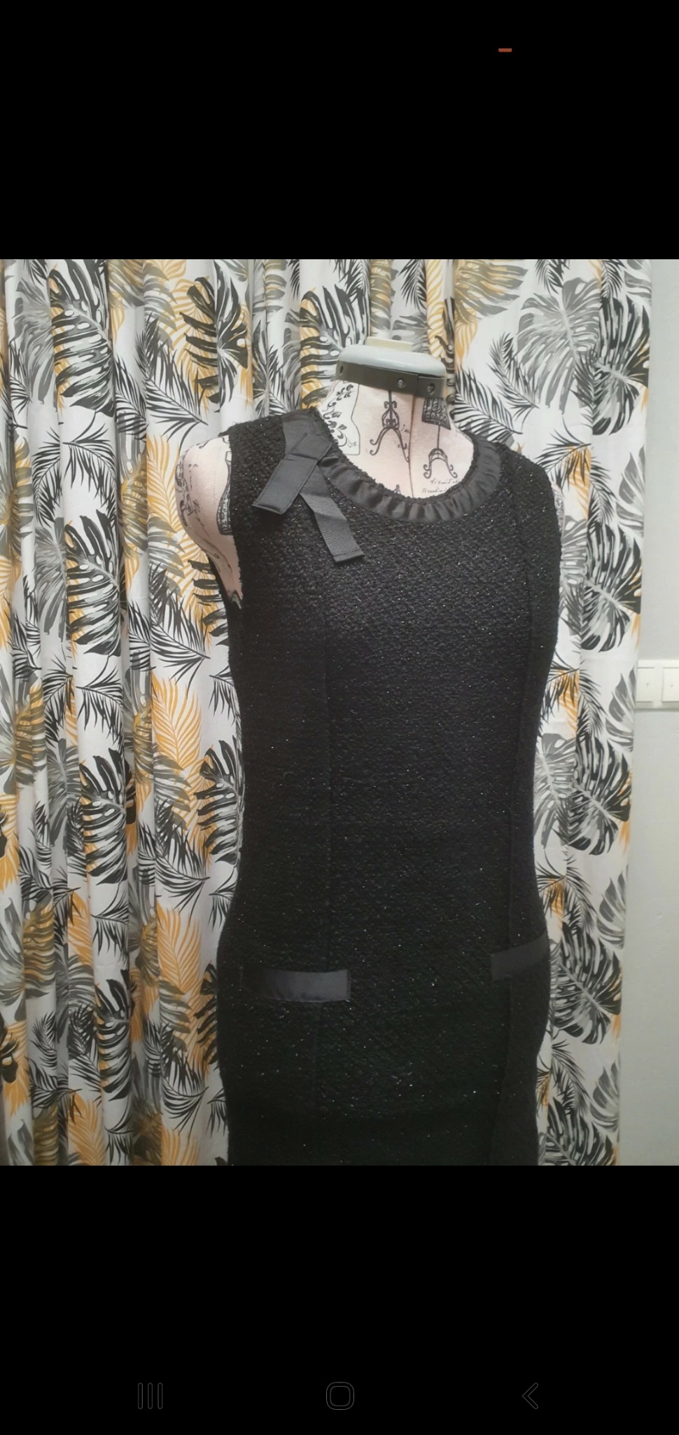 Czarna, tweedowa sukienka bez rękawów z satynowymi wykończeniami i kokardą na manekinie krawieckim na tle zasłony w liście.