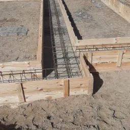 Przygotowanie szalunku i zbrojenia pod fundament z widocznym zbiornikiem na wodę w tle.