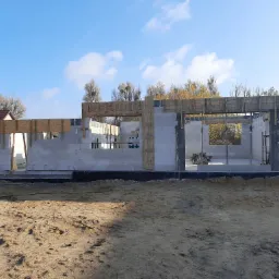 Wznoszenie ścian parterowego domu z bloczków betonowych, widoczne drewniane szalunki nad otworami okiennymi i drzwiowymi, teren budowy z piaskiem i fragmentami trawy, w tle domy jednorodzinne...