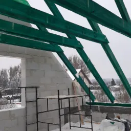 Wznoszenie ścian budynku z betonu komórkowego w zimowej scenerii, widoczne zielone elementy konstrukcji dachu i rusztowanie.