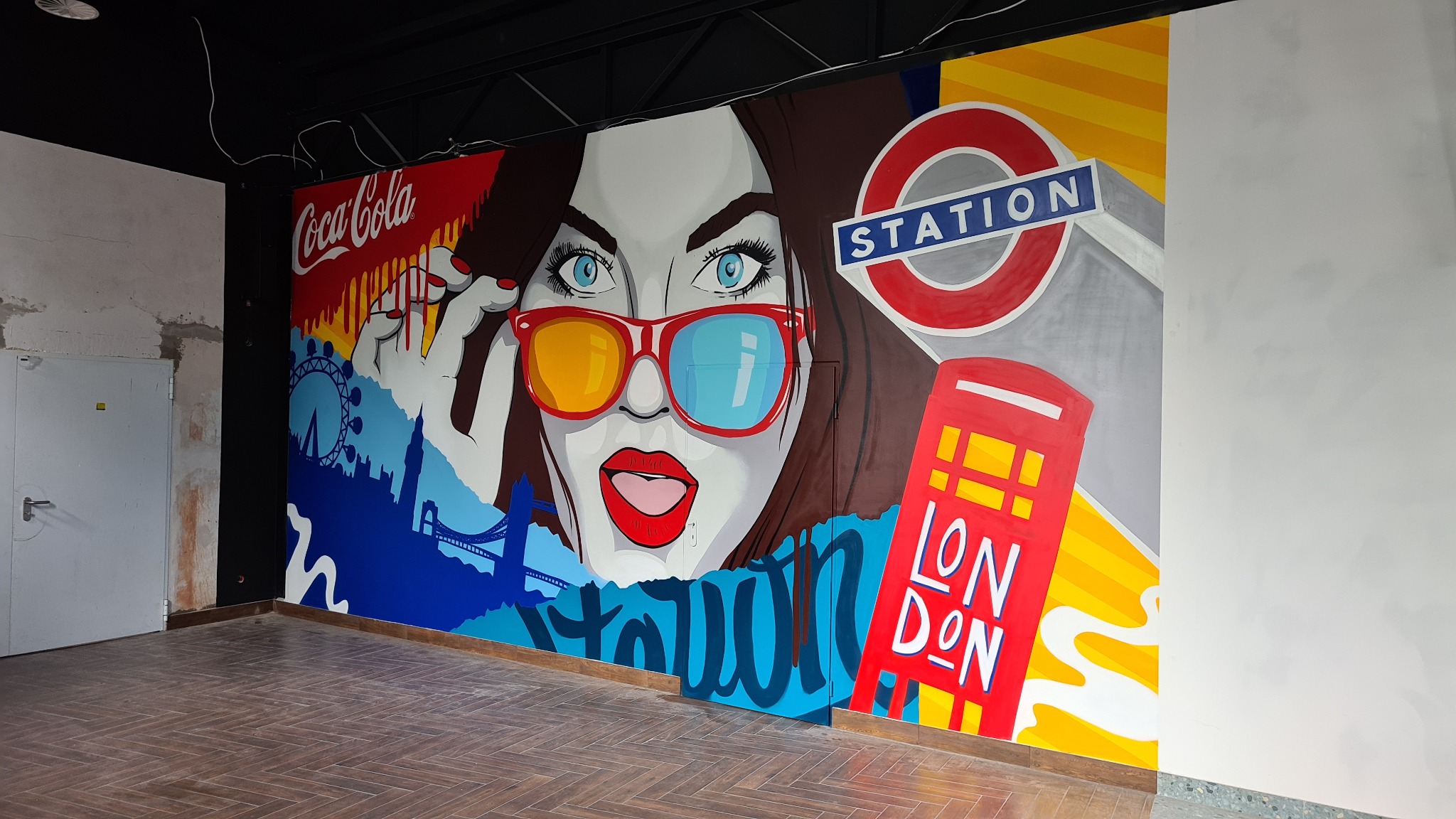 Pop-artowy mural z motywami Londynu: kobieta w okularach, budka telefoniczna, logo metra i panorama miasta, namalowany na ścianie w surowym wnętrzu.
