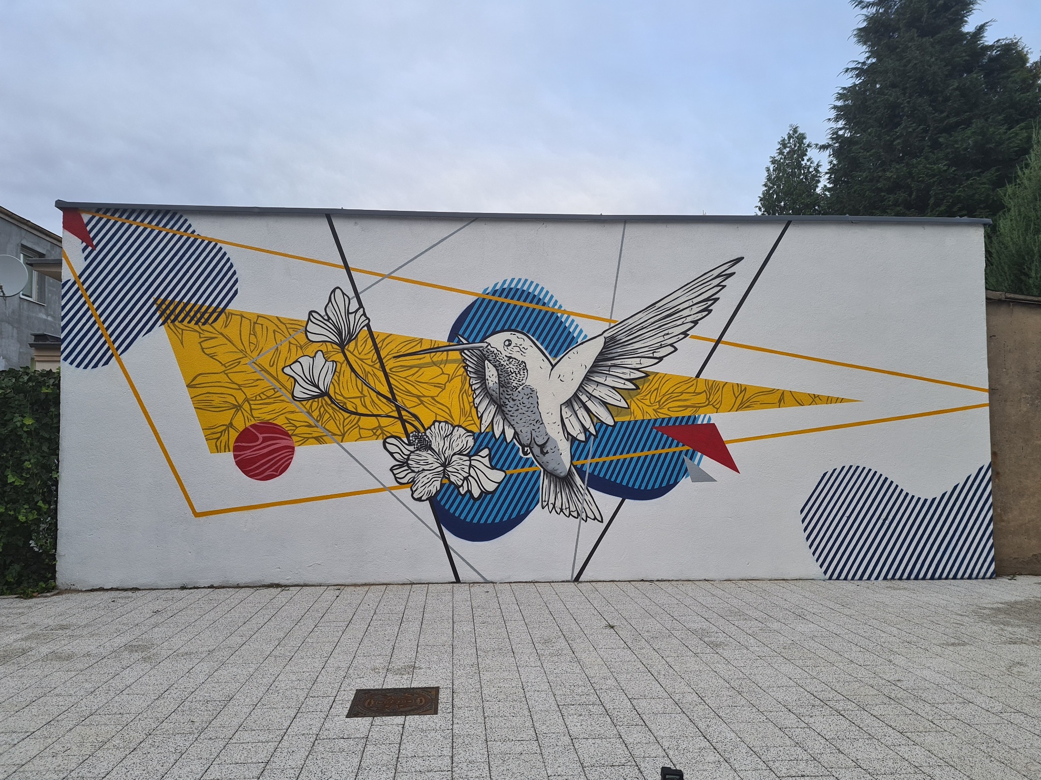 Ściana budynku ozdobiona muralem przedstawiającym kolibra w locie, otoczonego geometrycznymi kształtami, kwiatami i liśćmi na białym tle.