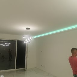 Paulina Rochmińska - Nowoczesne wnętrze z podświetlanym, zielonym paskiem LED na suficie, wiszącą lampą o spiralnym kształcie i marmurową podłogą, fotografowane przez mężczyznę telefonem.