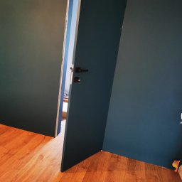 Paulina Rochmińska - Świeżo pomalowane na ciemny granat ściany i drewnopodobne panele podłogowe w mieszkaniu w Łodzi, widoczne otwarte drzwi i wystający fragment grzejnika z termostatem.