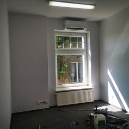 Paulina Rochmińska - Pomieszczenie po malowaniu na szaro, widoczne okno z widokiem na zieleń, grzejnik, klimatyzator, puszki z farbą i wałek malarski na podłodze.