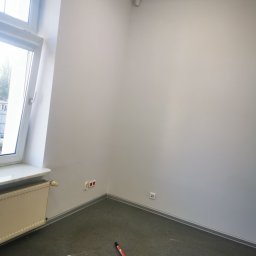 Paulina Rochmińska - Pusty pokój z pomalowanymi na biało ścianami, szarą podłogą, grzejnikiem pod oknem i widocznym fragmentem kija malarskiego na podłodze.
