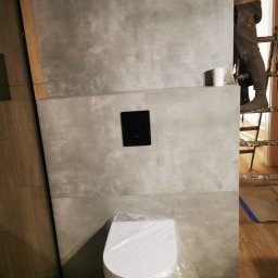 Paulina Rochmińska - Montaż szarej, wielkoformatowej płytki w łazience, widoczny fragment stelaża podtynkowego z czarnym przyciskiem spłukującym, zabezpieczona folią toaleta oraz pracownik na drabinie.