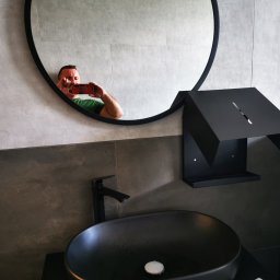 Paulina Rochmińska - Nowoczesna łazienka z czarną umywalką i okrągłym lustrem, w którym odbija się osoba robiąca zdjęcie telefonem, widoczny również dozownik na mydło w rękawicy ochronnej.