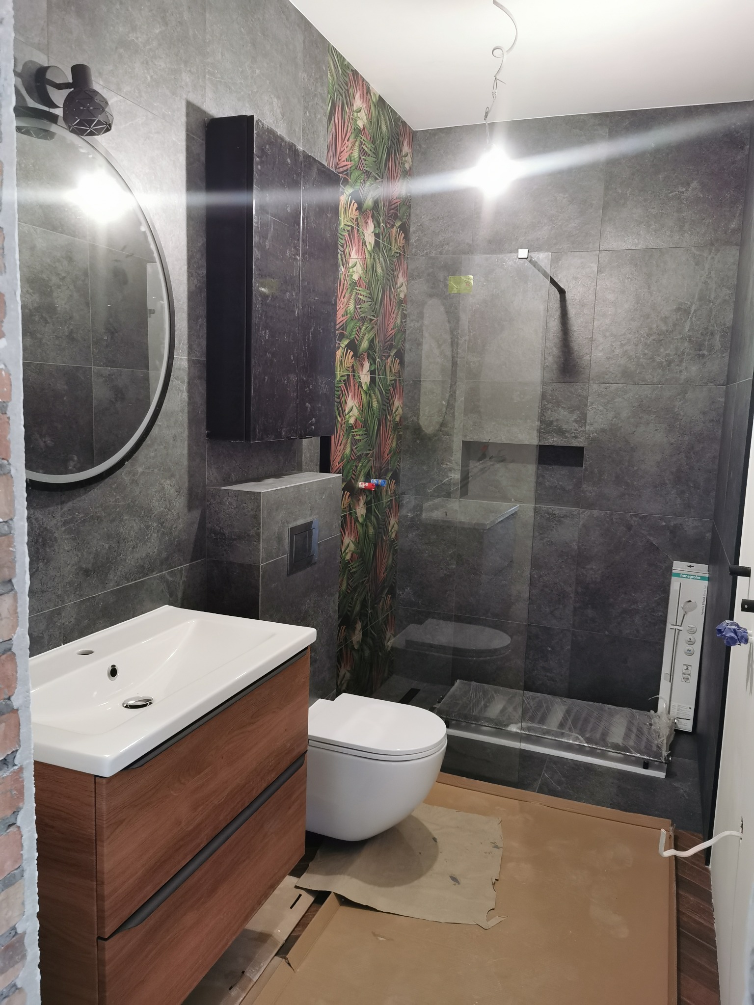 Wykończona łazienka z szarymi płytkami imitującymi kamień, drewnianą szafką pod umywalką, okrągłym lustrem z podświetleniem LED i kabiną prysznicową typu walk-in z tropikalnym akcentem na jednej ze...