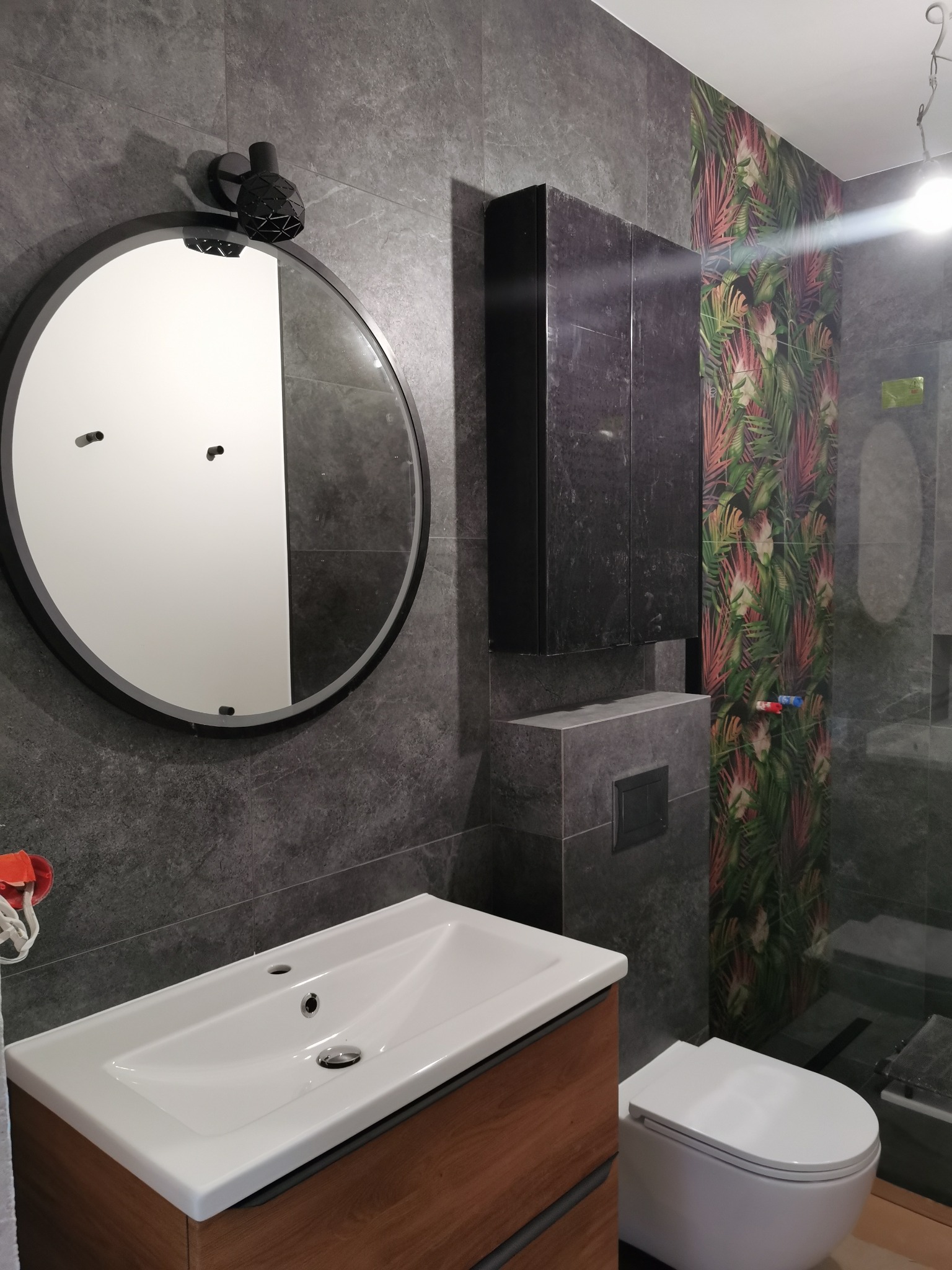 Nowoczesna łazienka z szarymi płytkami imitującymi kamień, okrągłym lustrem z podświetleniem, białą umywalką na drewnianej szafce, podwieszaną toaletą i szafką wiszącą, akcentem jest ściana...