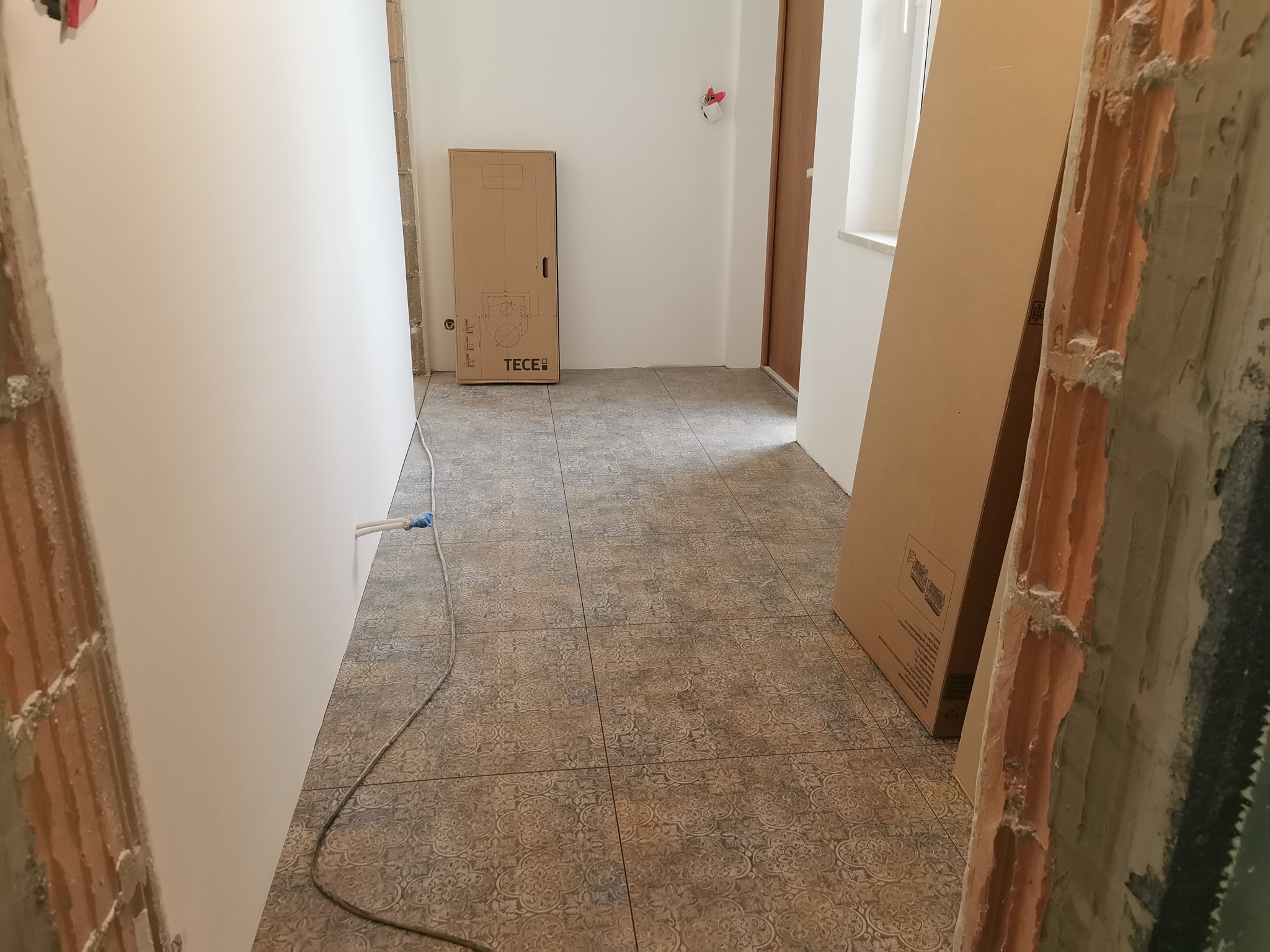 Wnętrze w trakcie remontu: świeżo położona podłoga z wzorzystych płytek, gładkie ściany, kartonowe opakowanie po sprzęcie sanitarnym marki TECE oraz surowe framugi z cegły.