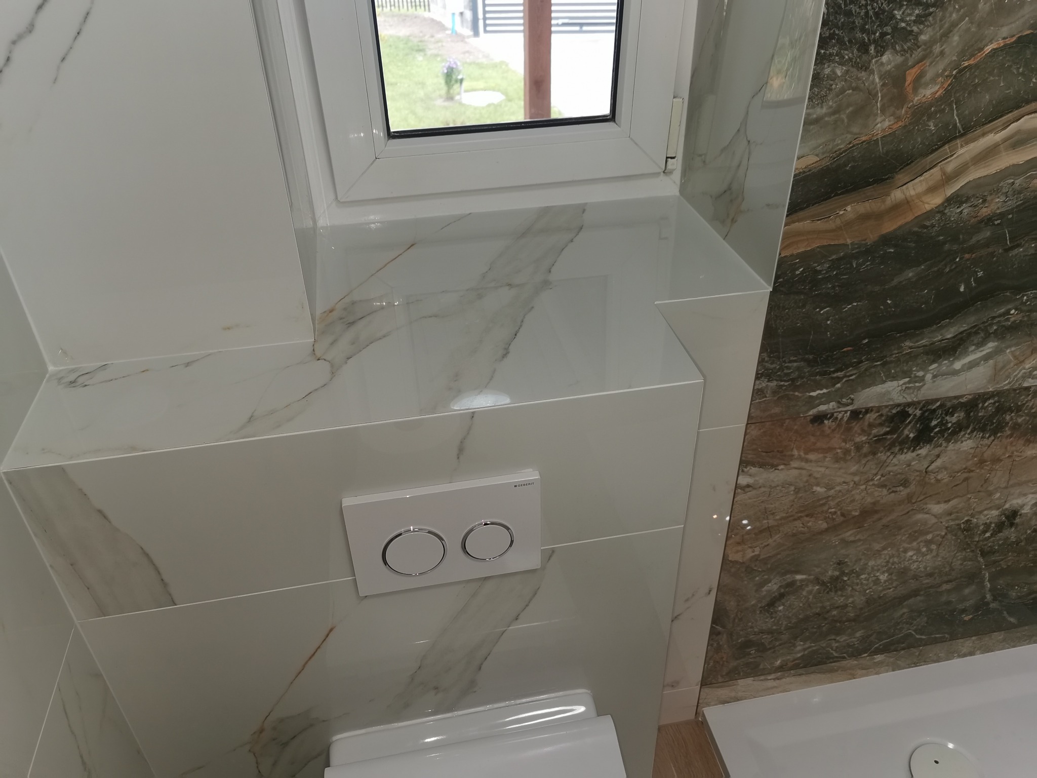 Nowoczesna łazienka z marmurowymi płytkami w odcieniach bieli i brązu, widoczny fragment toalety pod oknem i przycisk spłukiwania Geberit.