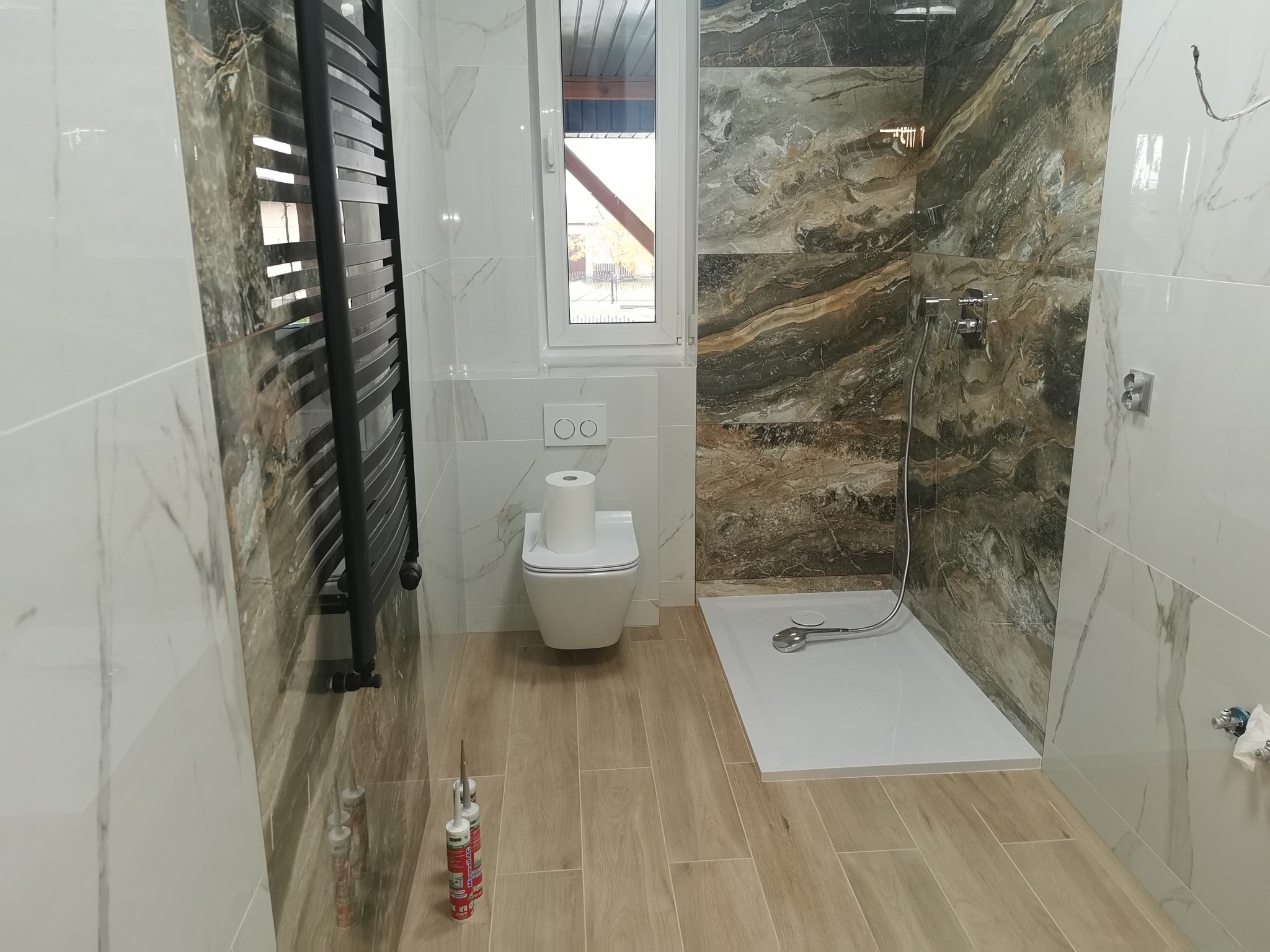 Wykończona łazienka z kabiną prysznicową, toaletą z papierem, grzejnikiem drabinkowym w kolorze czarnym, drewnopodobną podłogą i mieszanką płytek imitujących marmur w odcieniach szarości i bieli...