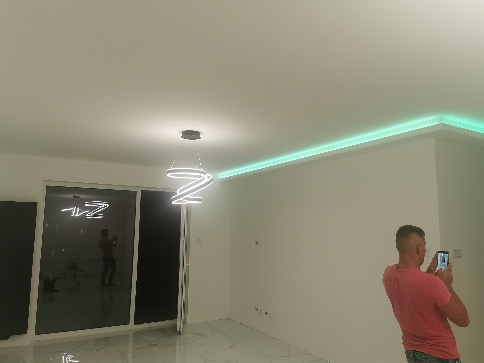 Nowoczesne wnętrze z podświetlanym, zielonym paskiem LED na suficie, wiszącą lampą o spiralnym kształcie i marmurową podłogą, fotografowane przez mężczyznę telefonem.