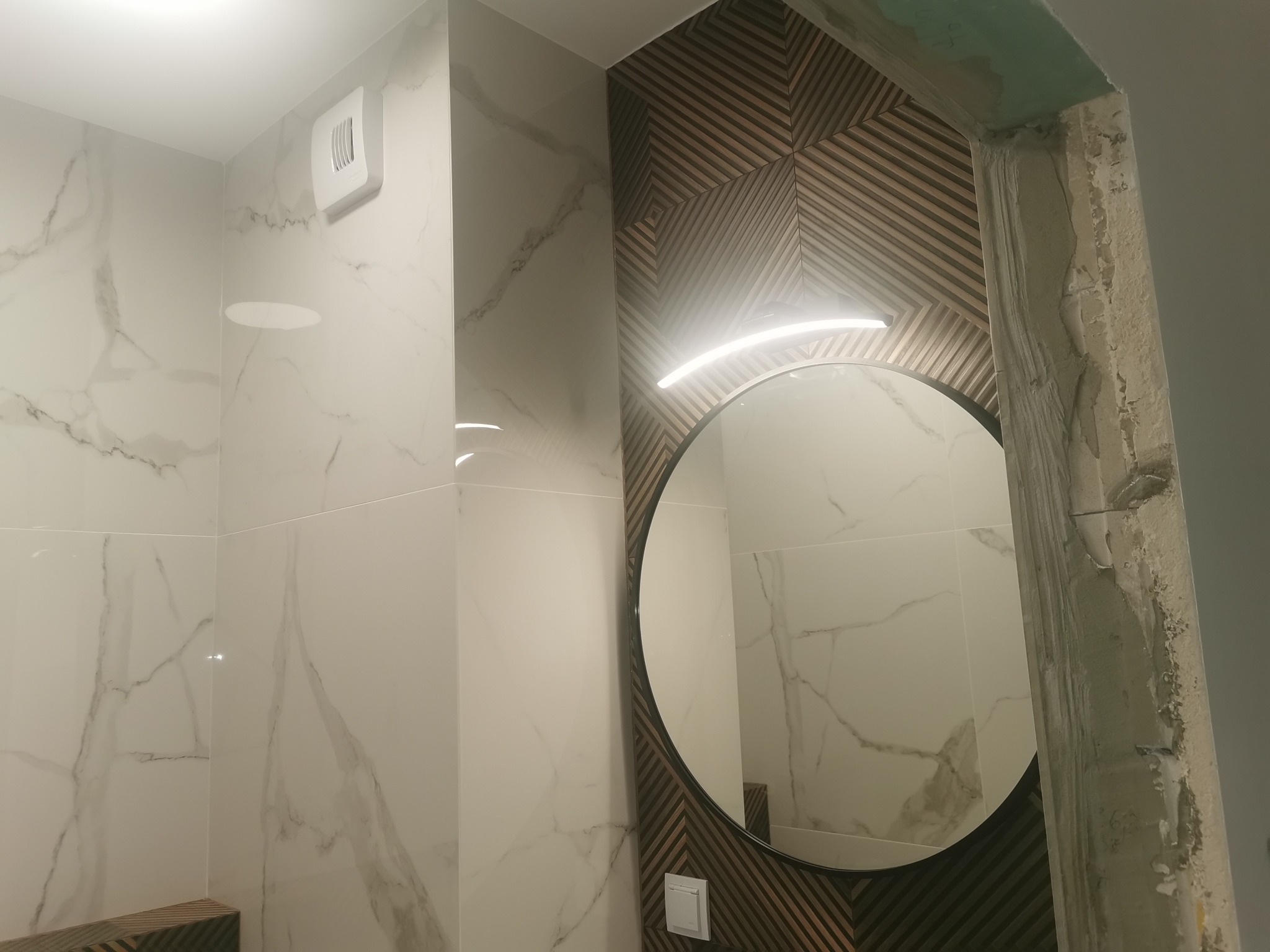 Nowoczesna łazienka z marmurowymi płytkami, okrągłym lustrem z podświetleniem LED i dekoracyjną ścianą z drewnianych paneli ułożonych w geometryczny wzór; widoczny fragment surowego wykończenia...