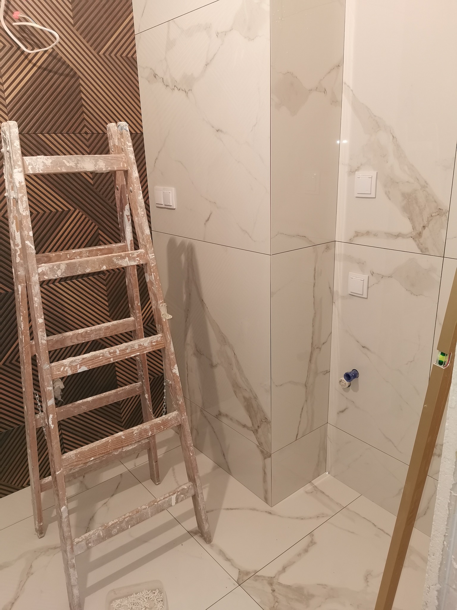 Remontowana łazienka z drewnianą drabiną, białe płytki imitujące marmur na ścianach i podłodze, dekoracyjna ściana z drewnianych paneli w tle, widoczne gniazdka i instalacja wodna.