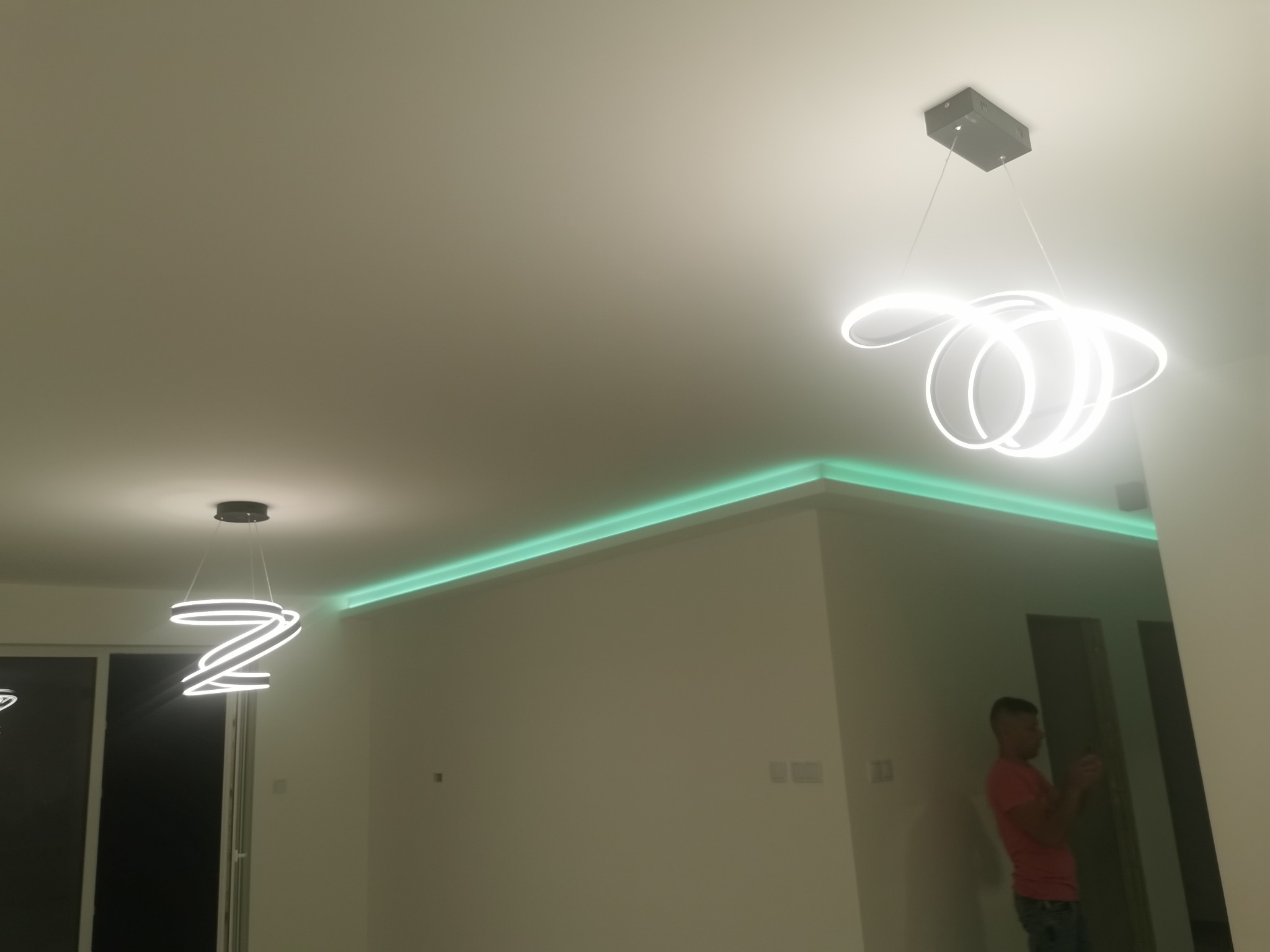 Nowoczesne, spiralne lampy LED wiszące na tle świeżo pomalowanego sufitu z zielonym oświetleniem LED wzdłuż krawędzi, widok z wnętrza mieszkania w Łodzi.