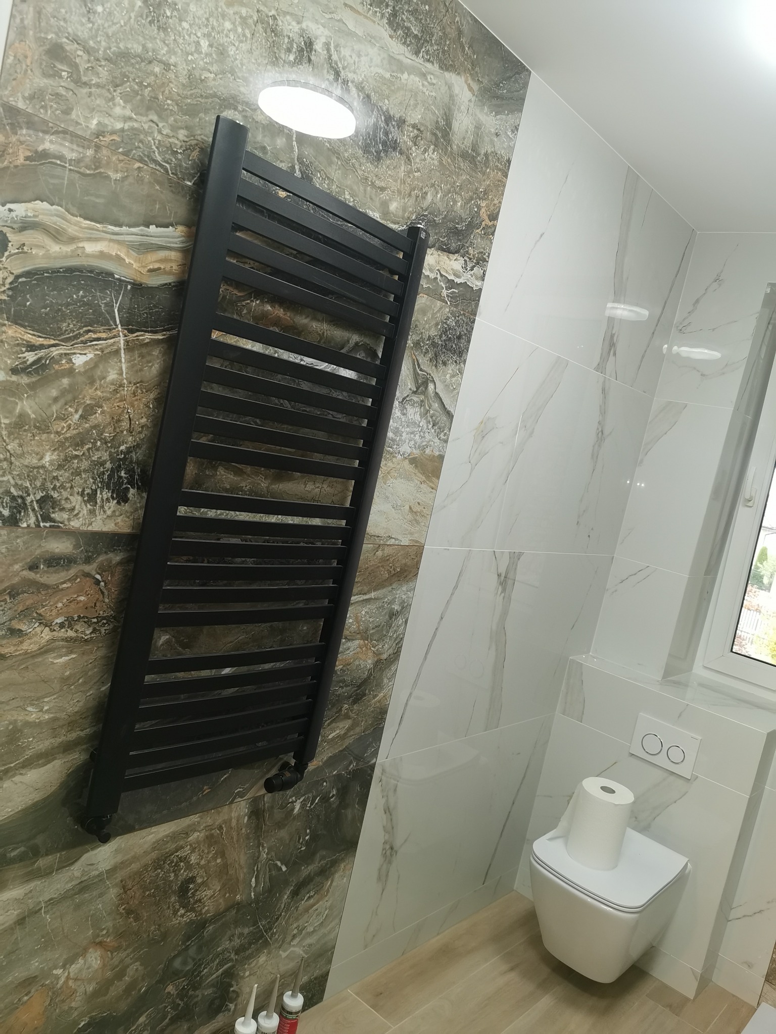 Nowoczesna łazienka z marmurowymi płytkami, czarnym grzejnikiem drabinkowym i podwieszanym WC z rolką papieru.