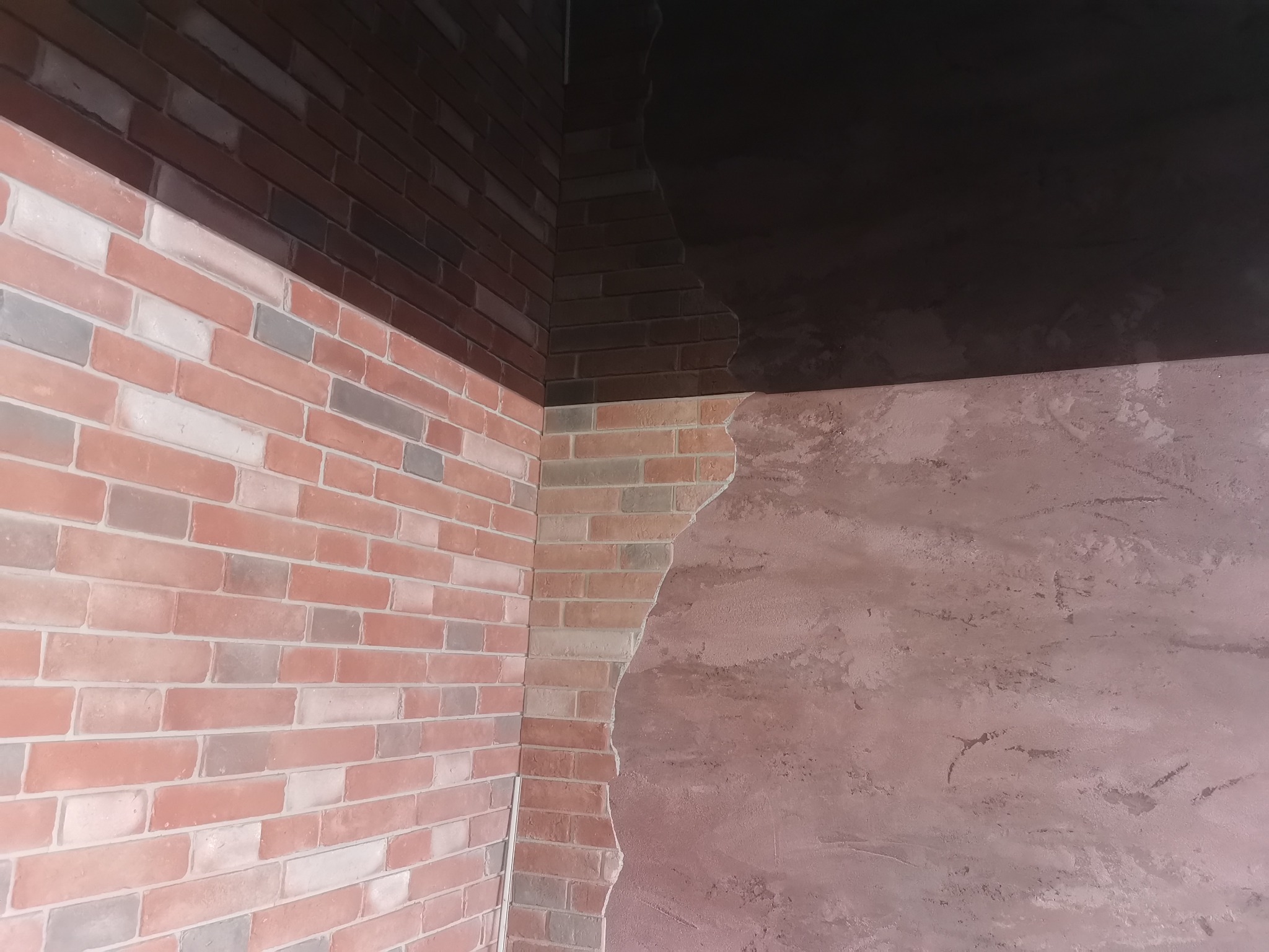 Fragment ściany z częściowo nałożoną czerwoną cegłą dekoracyjną i surową, różową fakturą, widoczny narożnik pomieszczenia i fragment ciemnego sufitu.