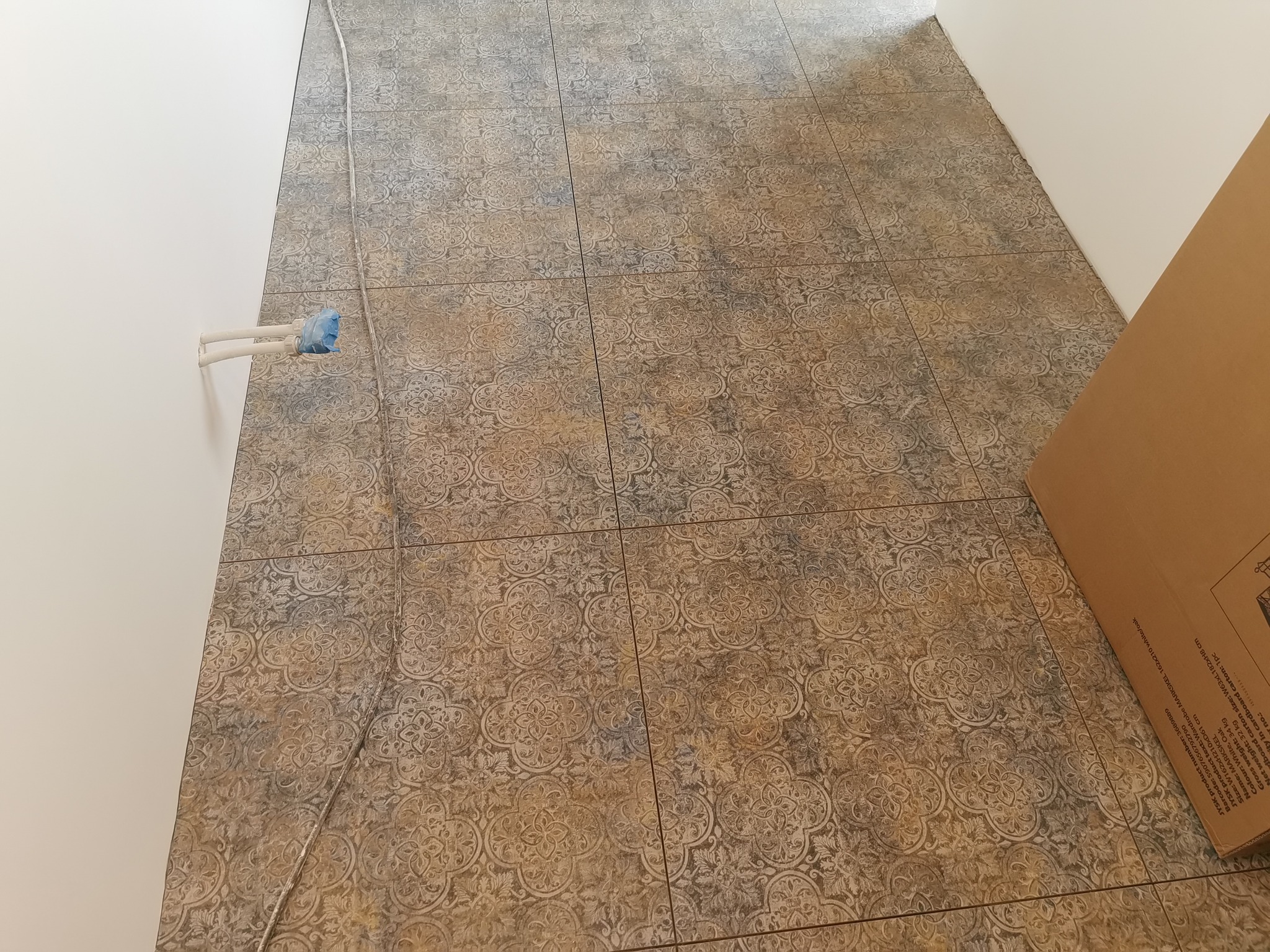 Ułożone płytki podłogowe z wzorem arabeski w odcieniach beżu i szarości, widoczne rury instalacji wodnej zabezpieczone folią oraz fragment kartonu w rogu kadru.