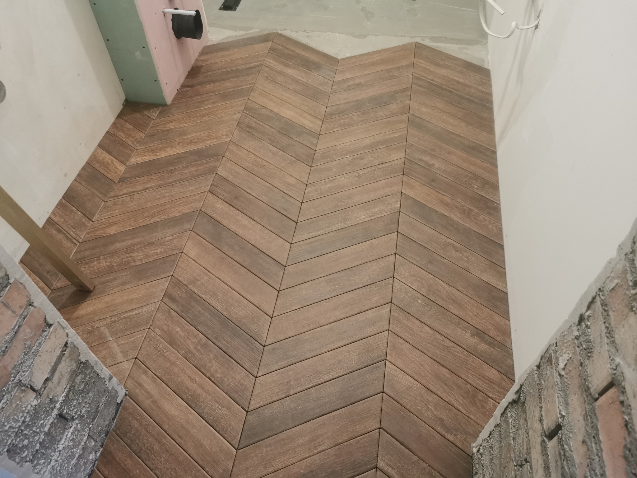 Ukończona podłoga z drewnopodobnych paneli ułożonych w jodełkę, widoczne fragmenty ścian w trakcie wykończenia, surowa ściana z cegły.