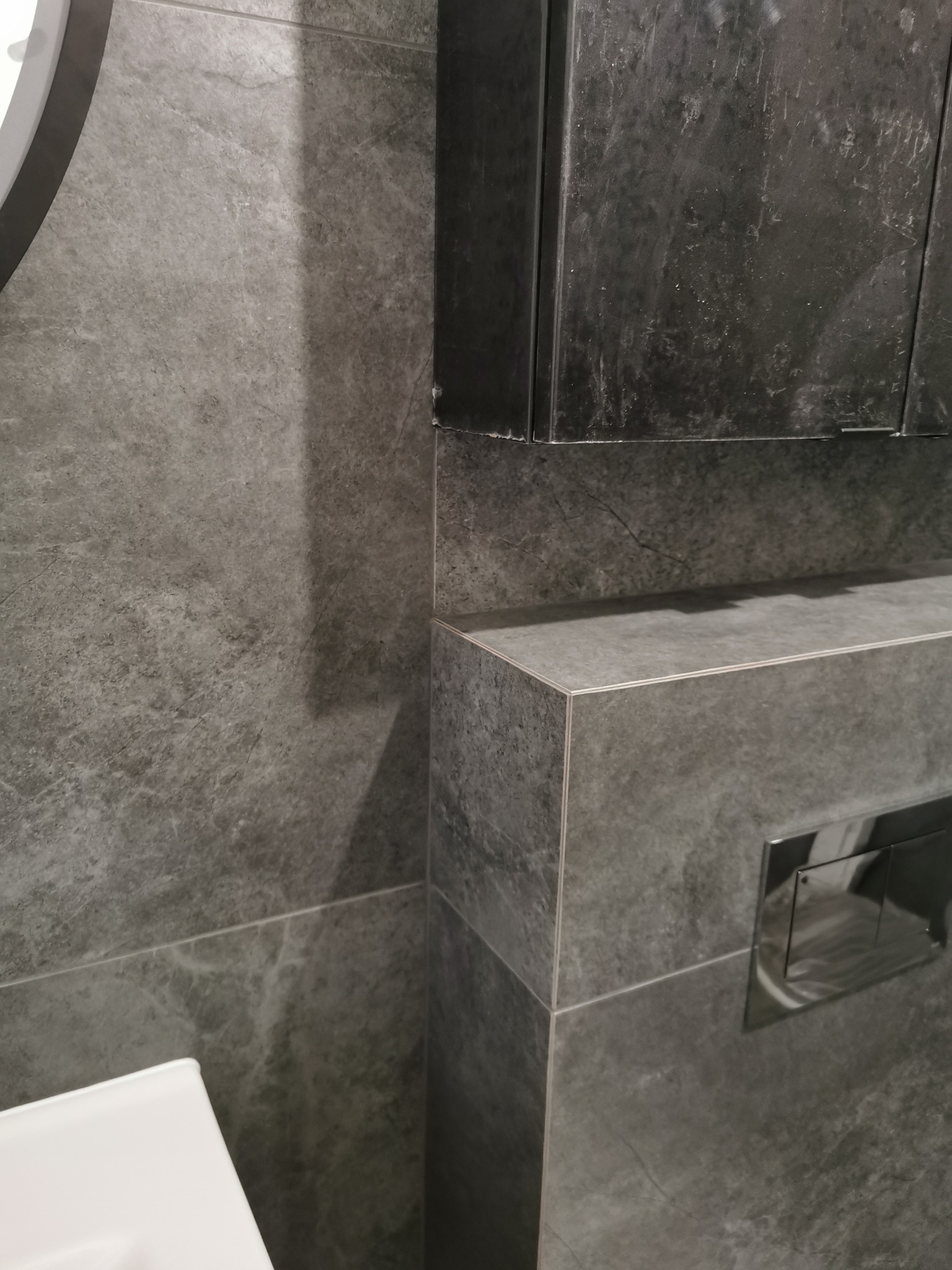 Szara łazienka z płytkami imitującymi kamień, zabudowana toaleta z chromowanym przyciskiem spłukiwania i szafka wisząca nad nią.