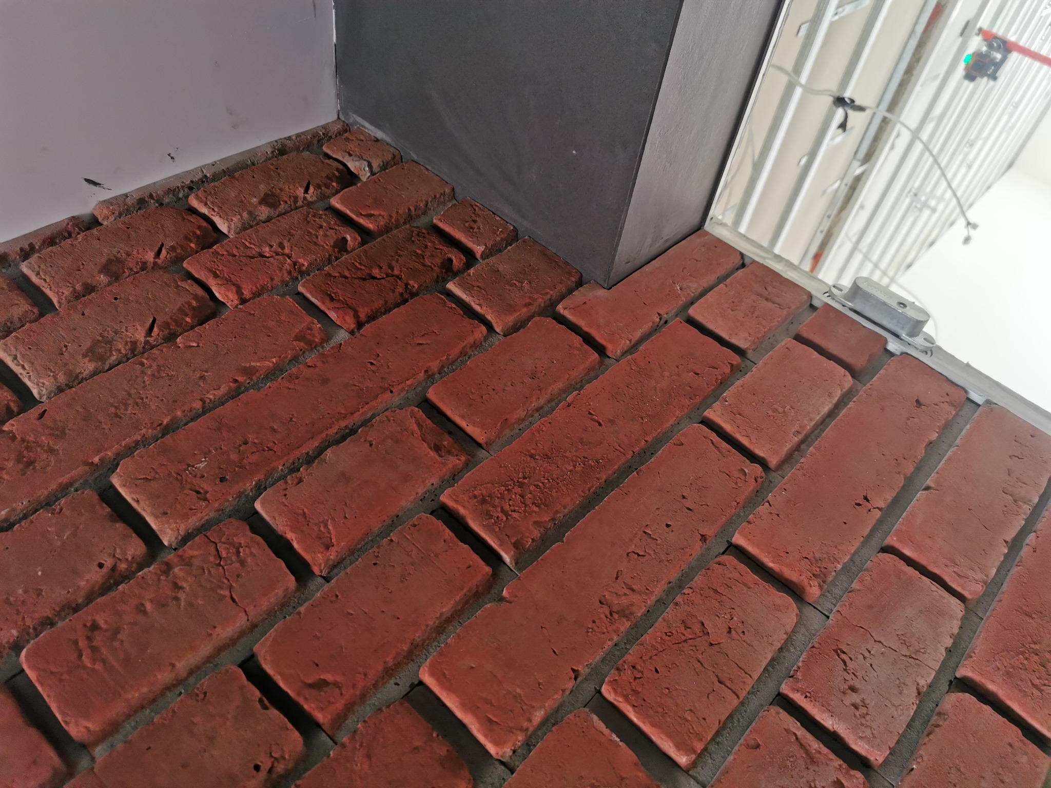 Czerwona ceglana okładzina ścienna z szarą listwą przypodłogową i widoczną konstrukcją sufitu podwieszanego w tle.