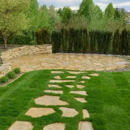 Kamienny taras z nieregularnych płyt, otoczony zielonym trawnikiem i żywopłotem z tui, prowadzący ścieżką z kamiennych płyt przez trawnik. Kamienna ścianka oporowa z nasadzeniami w tle.