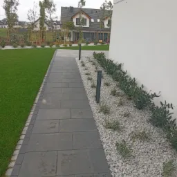 Nowoczesny ogród z szarą ścieżką z płyt, białym żwirem i niskimi roślinami, oświetlony lampami ogrodowymi, na tle domu z zielonym trawnikiem.