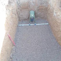 Przygotowanie podłoża pod taras: wykop z warstwą drenażową z grysu, pracownik wyrównuje poziom za pomocą poziomicy, widoczne ściany wykopu i narzędzia.