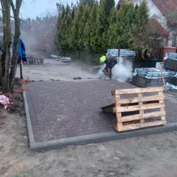 Pracownik w kasku tnący kostkę brukową na posesji z częściowo ułożonym brukiem, paletą i stertami kostki w tle.