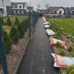 Przygotowanie terenu pod trawnik: rozłożona czarna mata i worki z korą sosnową wzdłuż ogrodzenia posesji z nowymi domami w tle.