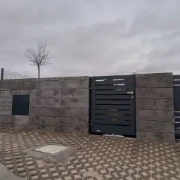 Nowoczesne ogrodzenie z szarych betonowych bloków i metalowa brama z poziomymi szczeblami, uzupełnione ażurową kostką brukową i młodym drzewkiem bez liści na tle pochmurnego nieba.