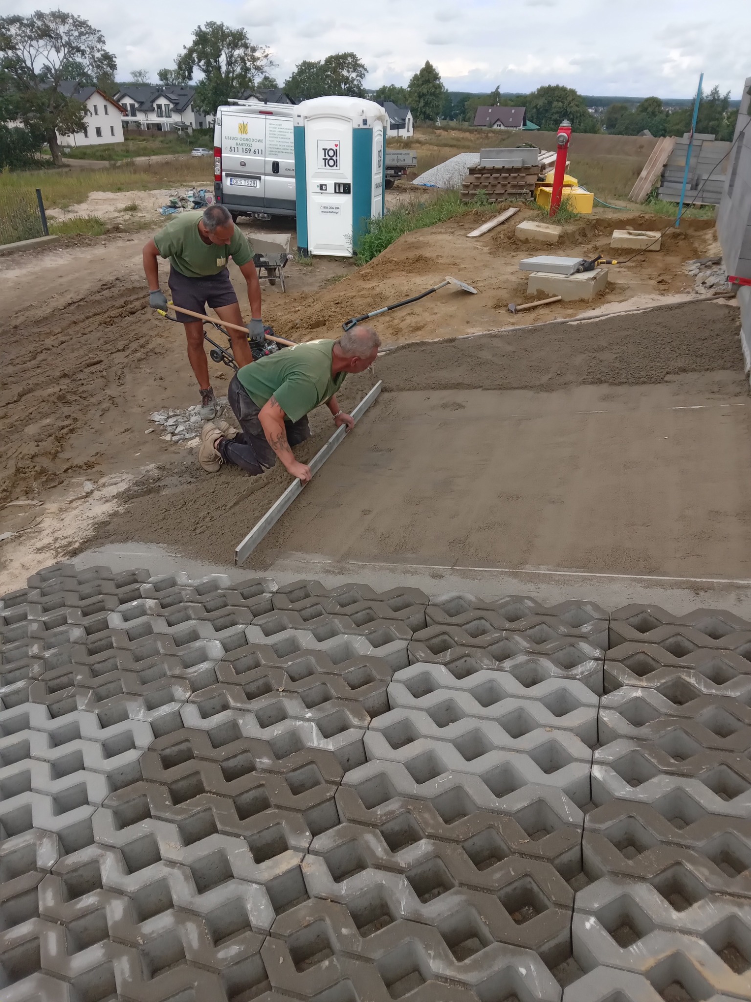 Dwóch mężczyzn w trakcie prac przygotowawczych pod ułożenie ażurowych płyt chodnikowych, jeden wyrównuje podłoże, drugi zagęszcza ziemię, w tle widoczny samochód dostawczy i przenośna toaleta.