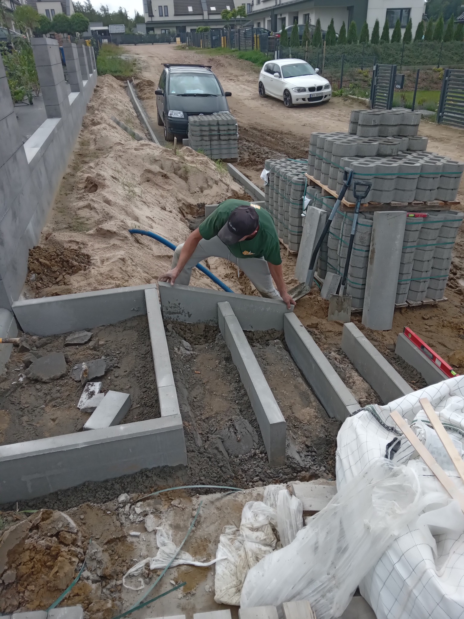 Pracownik w Kościerzynie układa fundament z betonowych bloczków na posesji, widoczne narzędzia i palety z kostką brukową w tle, przygotowania do budowy ogrodzenia.