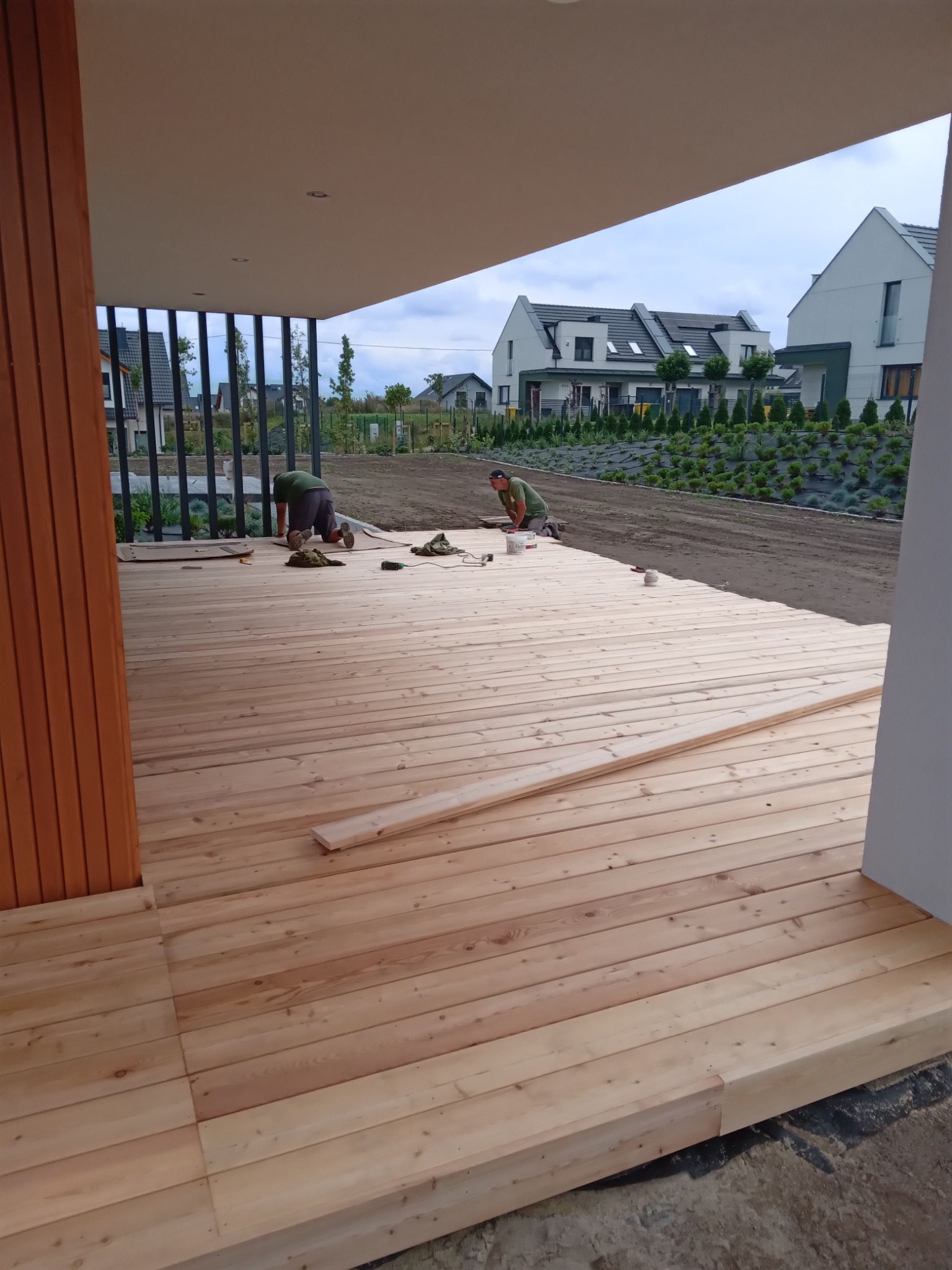 Montaż desek na drewnianym tarasie w trakcie realizacji, dwóch pracowników w tle, widok na ogród i zabudowę mieszkalną.