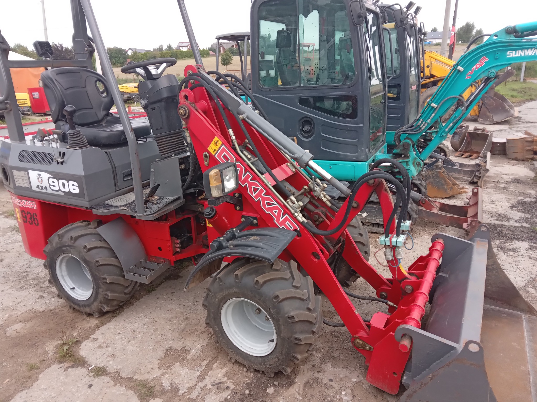 Czerwona ładowarka przegubowa DAKKAR 906 na placu budowy z innymi maszynami w tle.