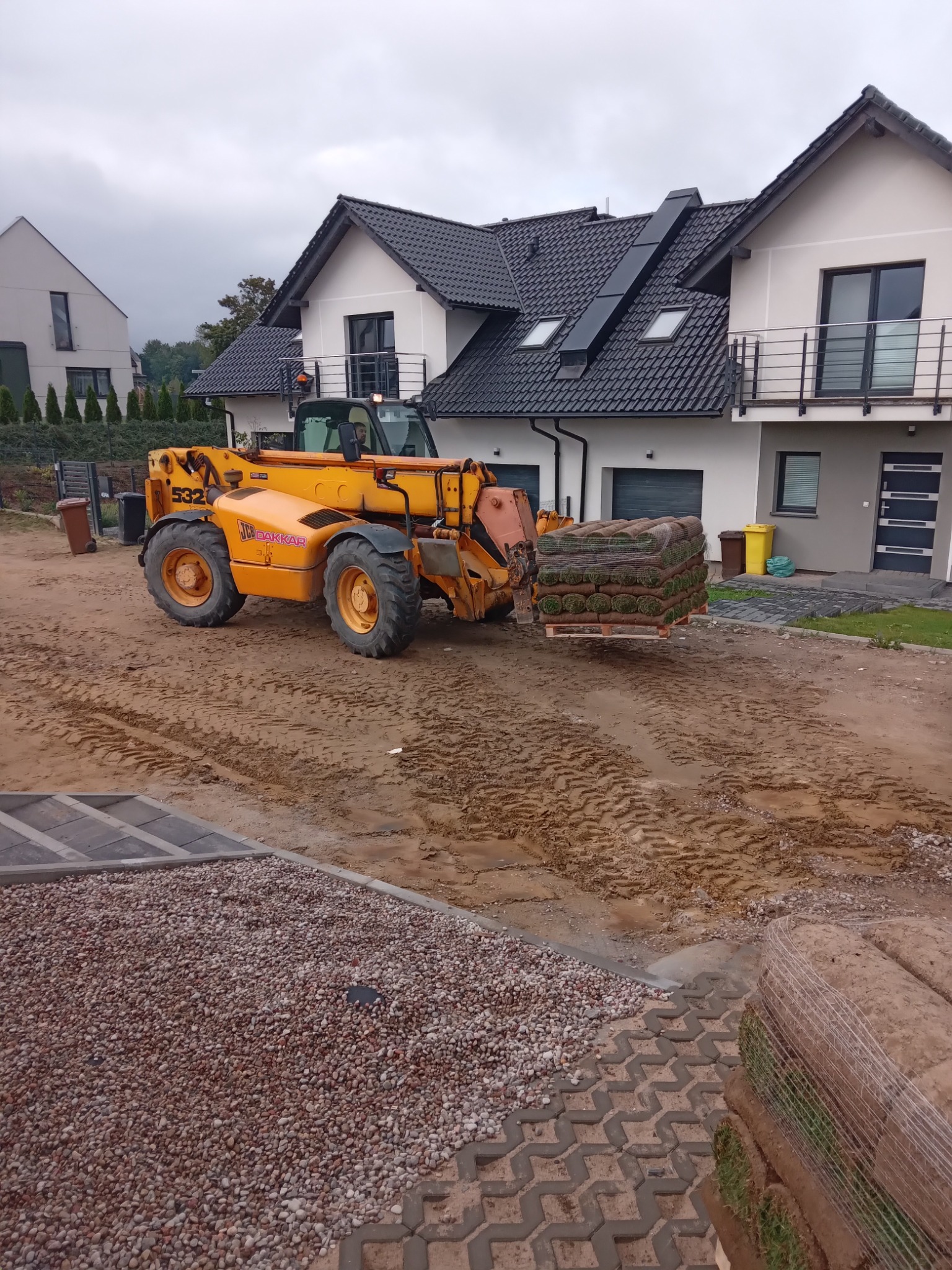 Żółta ładowarka teleskopowa przewozi paletę z rolkami trawy na tle nowoczesnego domu z szarą elewacją, widoczne ślady opon na ziemi i przygotowane podłoże z kamyczków.