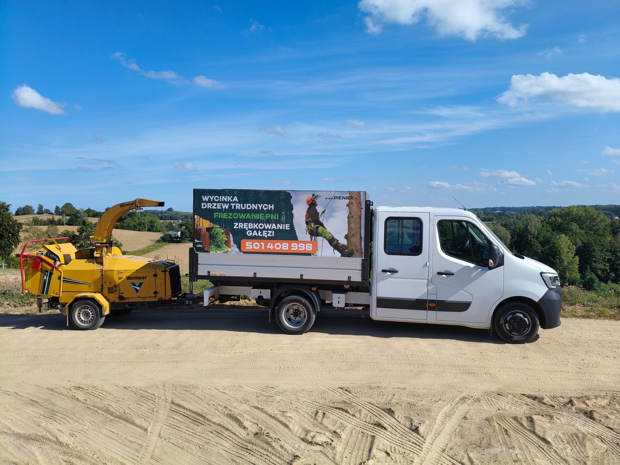 Żółty rębak do gałęzi Vermeer podczepiony do białego samochodu dostawczego z reklamą usług wycinki drzew trudnych i frezowania pni na tle błękitnego nieba i zieleni drzew.