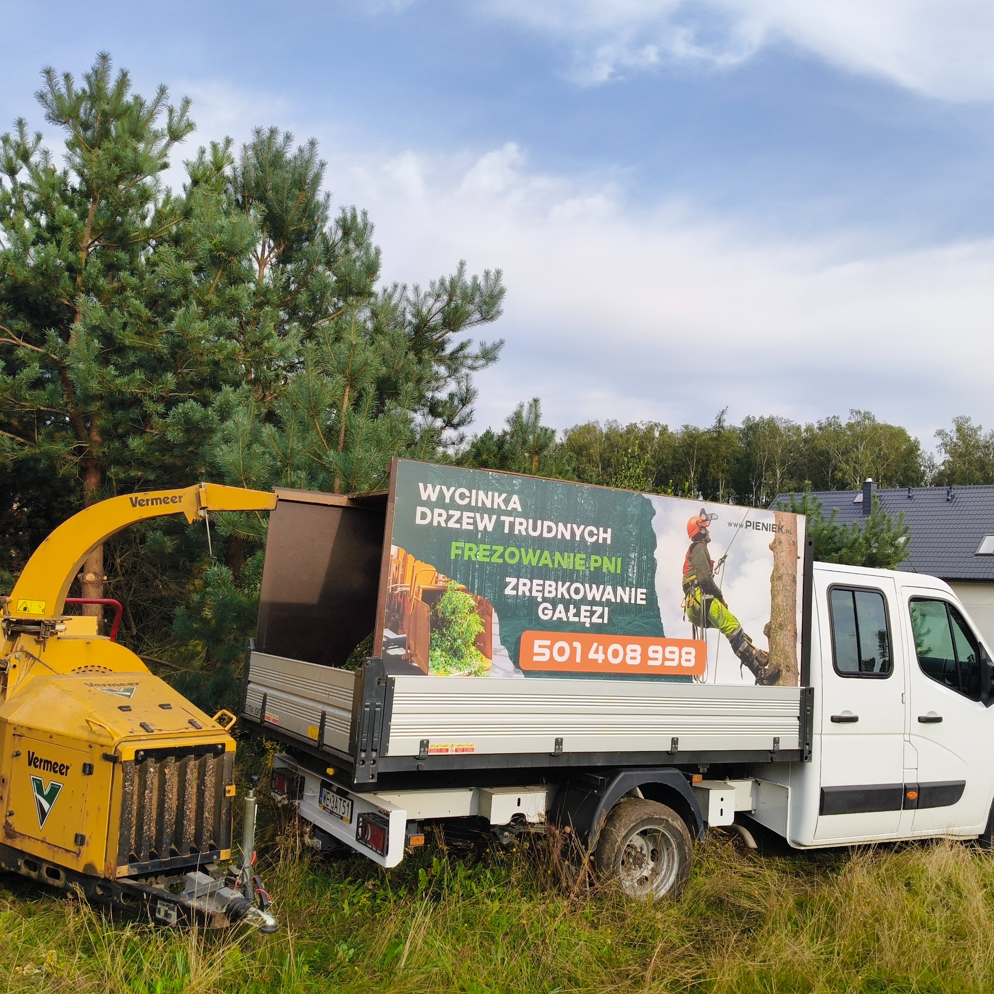 Żółty rębak Vermeer obok białego samochodu dostawczego z reklamą usług związanych z drzewami, w tle drzewa iglaste i fragment dachu domu.