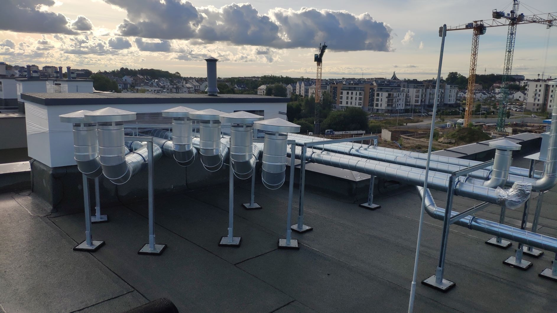 System wentylacji z metalowymi rurami na dachu budynku, widoczne kominki wentylacyjne z daszkami, w tle zabudowa miejska i żurawie budowlane.