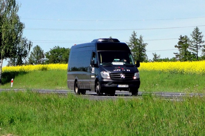 Czarny bus Mercedes-Benz z klimatyzacją na dachu, jadący po asfalcie na tle łąki z żółtymi kwiatami i drzewami. Widoczny numer rejestracyjny i logo firmy.