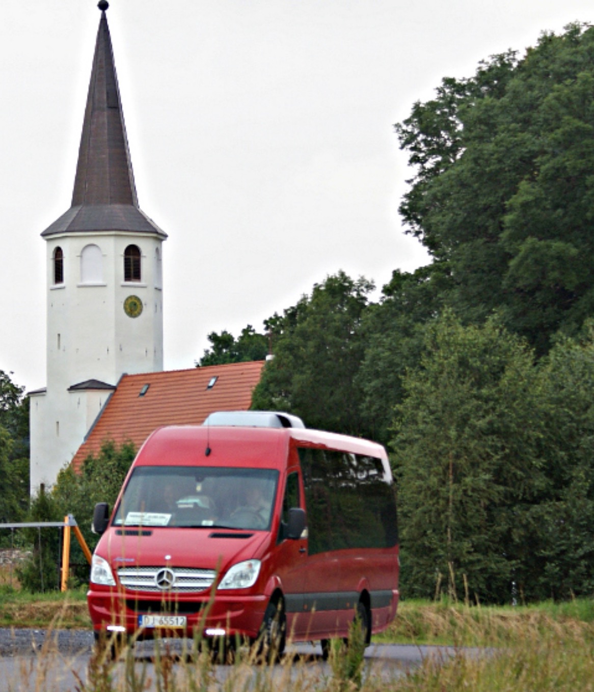 Czerwony bus Mercedes-Benz na tle wiejskiego kościoła z wieżą zegarową, otoczonego zielenią. Transport osobowy w malowniczej scenerii.